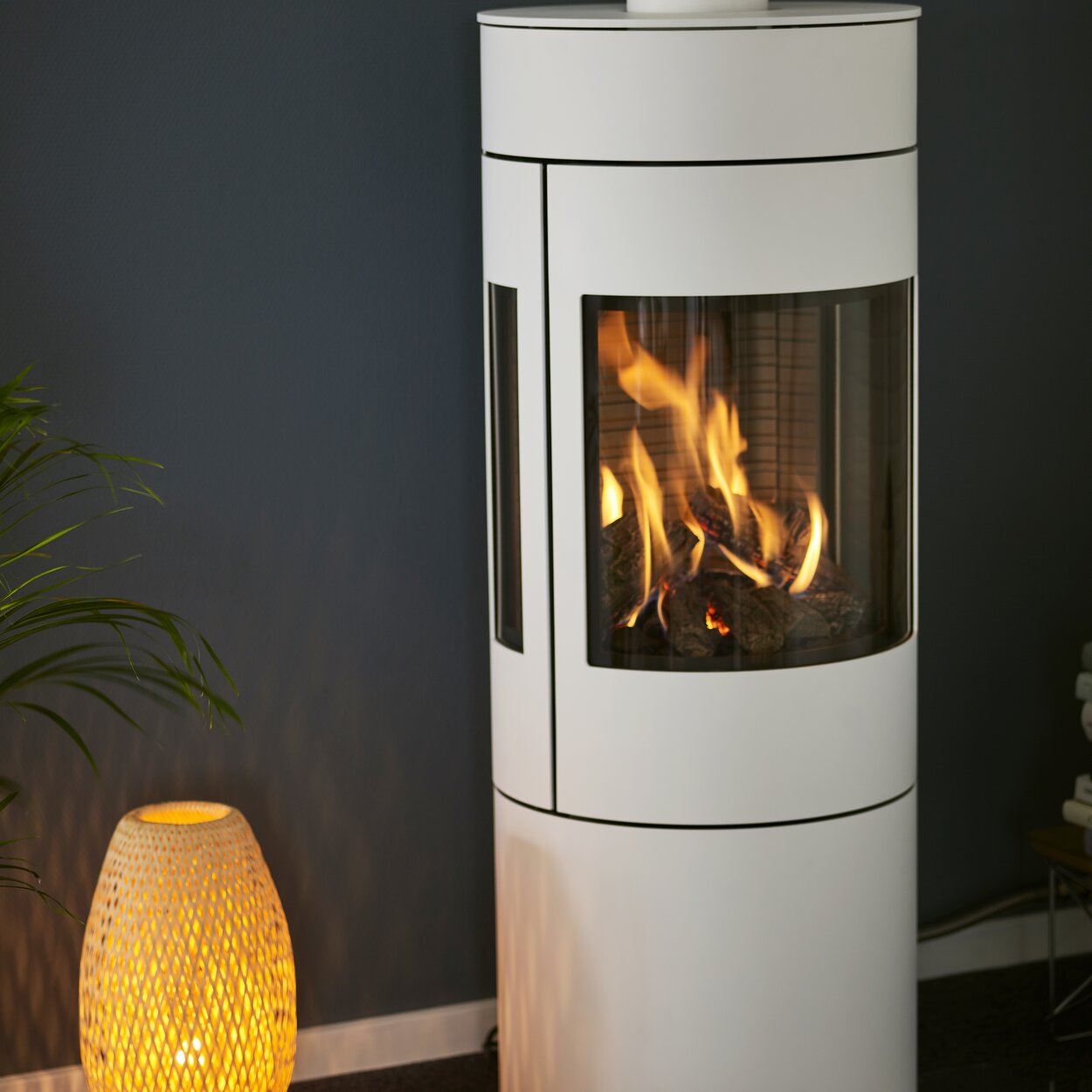 Gas stove VIVA 120 L in white with steel door and side windows in front of a dark blue wall next to a cosy floor lamp