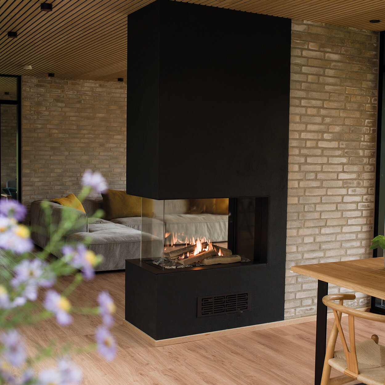 Gas-Kamin VISIO 70 RD steht als Raumteiler in einer schwarzen Trennwand und strukturiert so den eleganten Wohnraum mit Holzboden zwischen Wohnzimmer und Esstisch