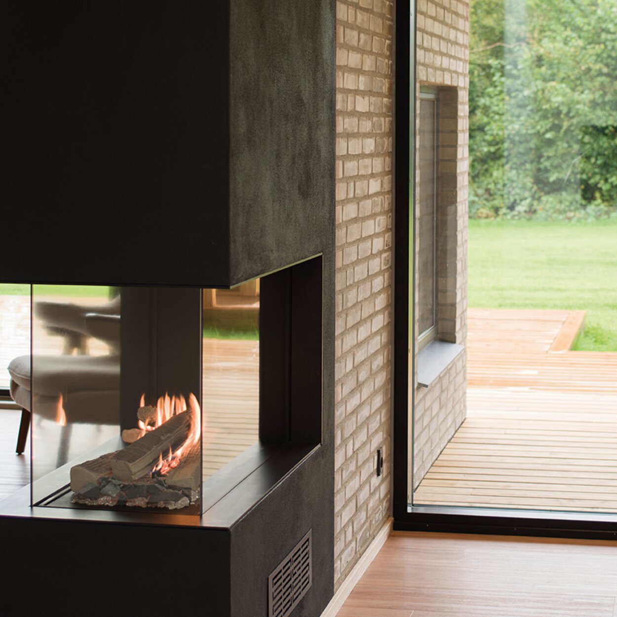 La cheminée à gaz VISIO 70 RD se dresse comme un séparateur de pièce dans une cloison noire, structurant ainsi l'élégante pièce à vivre avec son plancher en bois et ses grandes portes-fenêtres donnant sur le jardin