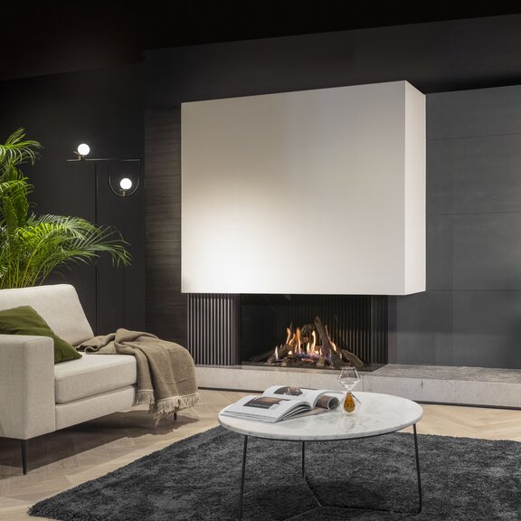 Camino a gas Gi1110/55C nel soggiorno con rivestimento bianco e grigio, divano grigio con cuscini e copriletto, pavimento in legno con tappeto nero