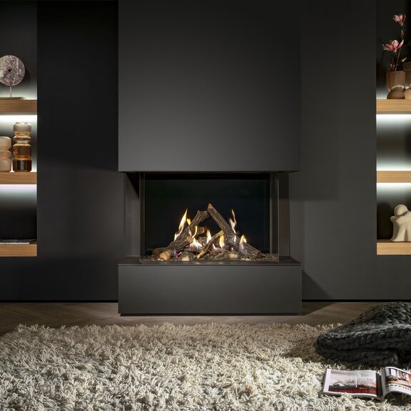 Camino a gas Gi85/55S con vetratura su 3 lati nel soggiorno, con parete nera, mensole in legno con elementi decorativi, tappeto bianco e coperta di lana sul pavimento