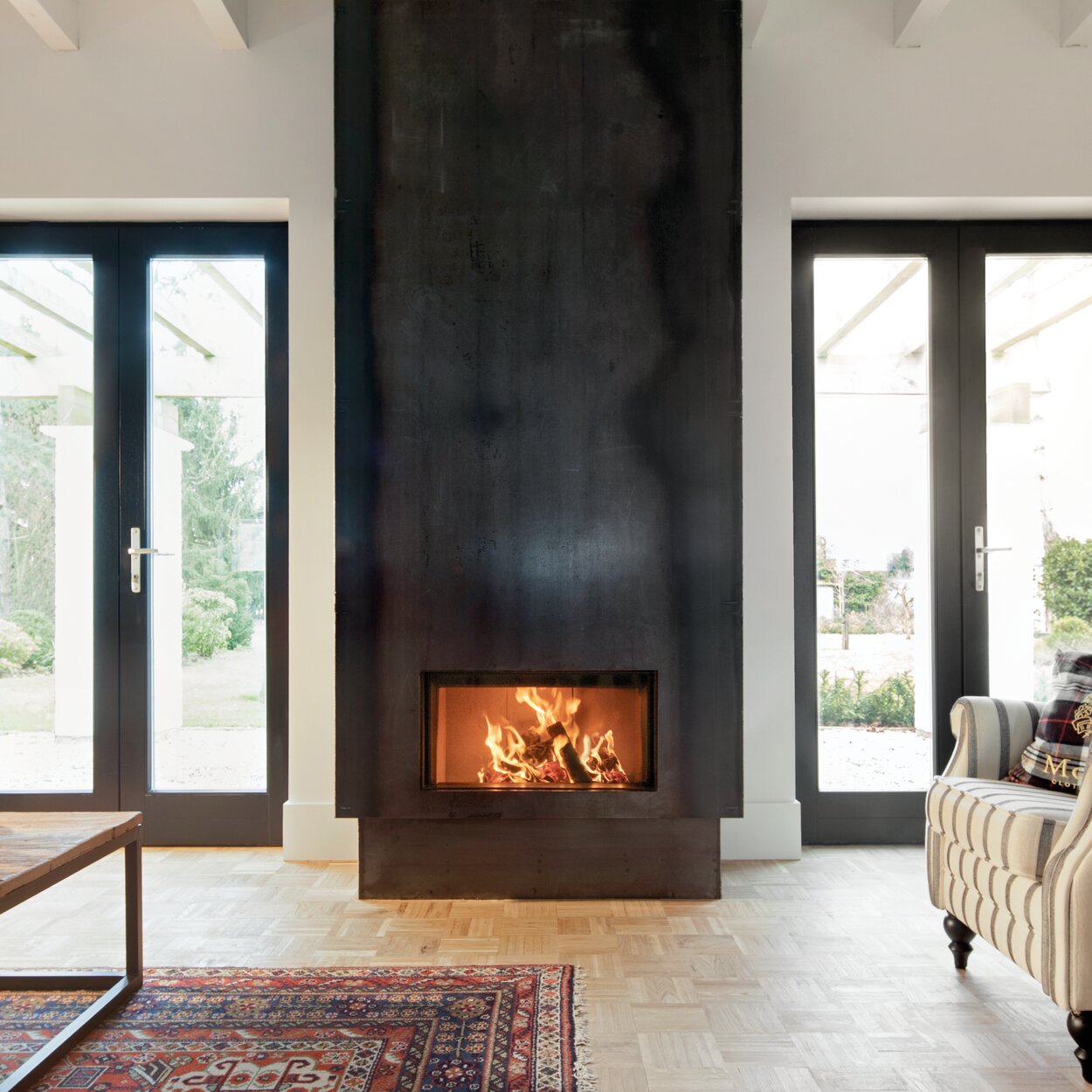 Cheminée à bois W70/33F de Kalfire avec revêtement en acier noir dans un salon clair, centré entre deux grandes portes-fenêtres